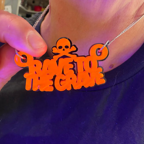 A close-up of a person wearing a silver chain necklace with a bright orange and black 'RAVE TO THE GRAVE' skull and crossbones pendant, held by a hand.
