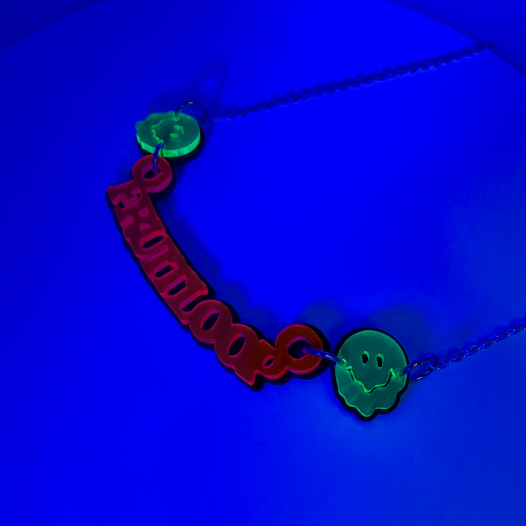 A necklace featuring a red 'fruitloop' cutout pendant and two glowing green melty face smiley charms on a dark blue background.