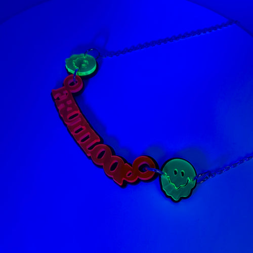 A necklace featuring a red 'fruitloop' cutout pendant and two glowing green melty face smiley charms on a dark blue background.