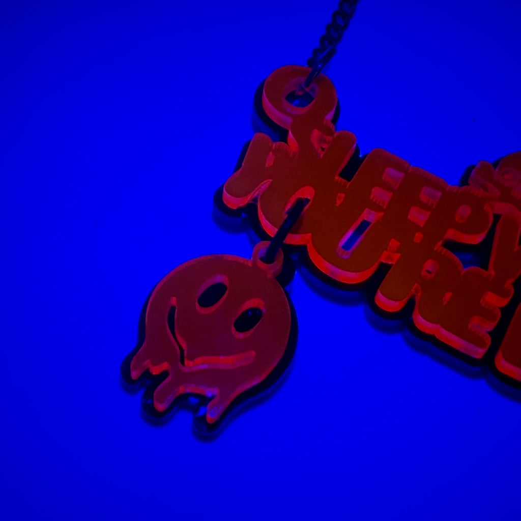 UV-reactive acrylic pendant necklace glowing red under blue light, featuring a ghost charm and stylized text, shown against a dark blue background.