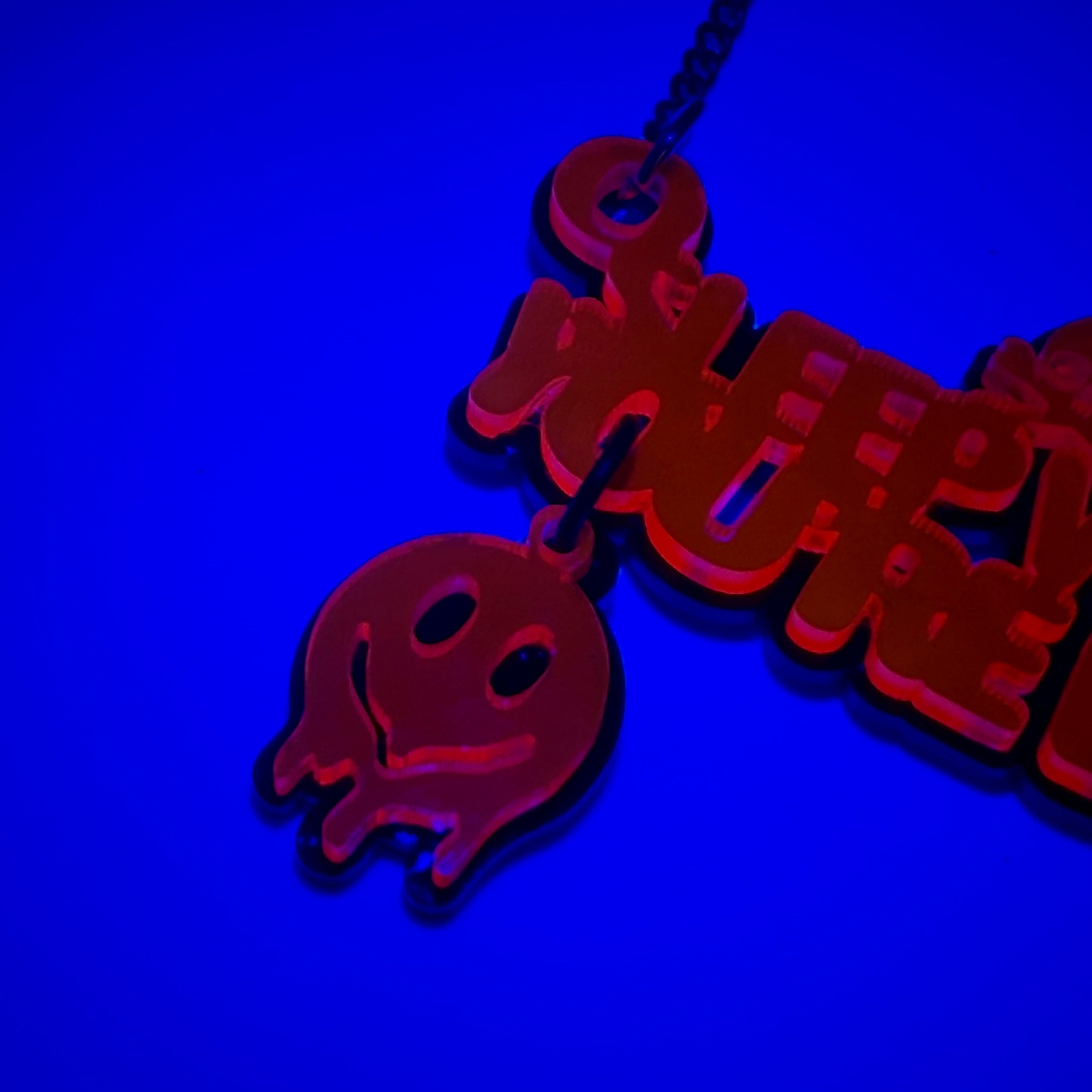 UV-reactive acrylic pendant necklace glowing red under blue light, featuring a ghost charm and stylized text, shown against a dark blue background.