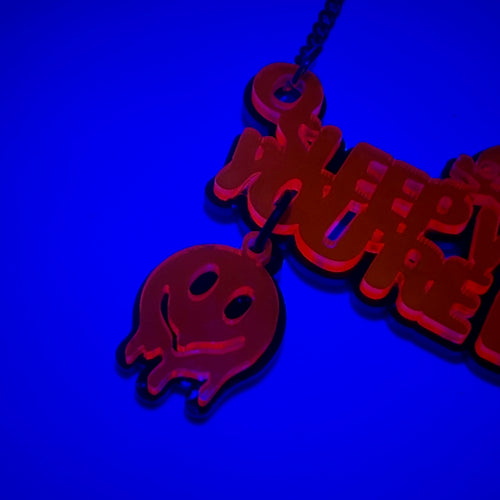UV-reactive acrylic pendant necklace glowing red under blue light, featuring a ghost charm and stylized text, shown against a dark blue background.