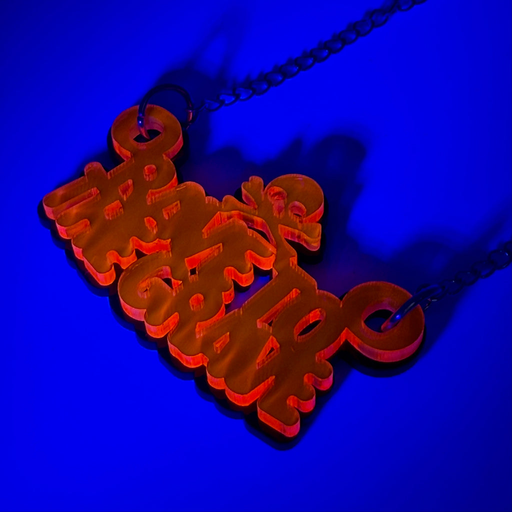 A neon orange acrylic pendant necklace spelling 'rave to the grave' in stylized script, hanging from a dark chain against a vibrant blue background.