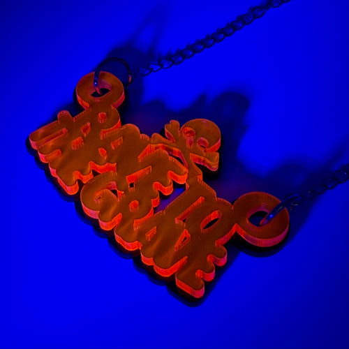 A neon orange acrylic pendant necklace spelling 'rave to the grave' in stylized script, hanging from a dark chain against a vibrant blue background.
