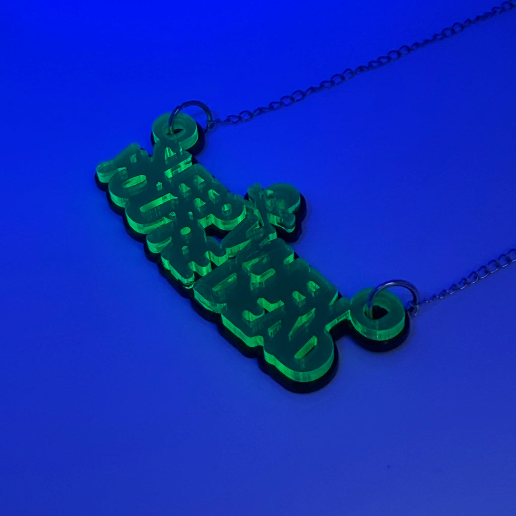 A green acrylic pendant necklace spelling 'Sleep when you're dead' in a stylized font, hanging from a silver chain against a solid blue background.