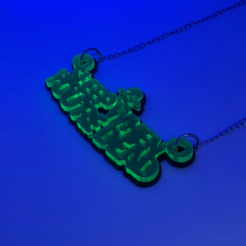 A green acrylic pendant necklace spelling 'Sleep when you're dead' in a stylized font, hanging from a silver chain against a solid blue background.