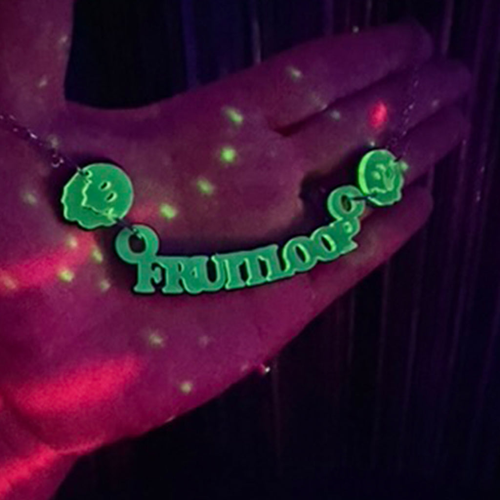 A blacklight-reactive necklace spelling 'fruitloop' with two green smiley faces, shown worn on a hand under UV lighting.