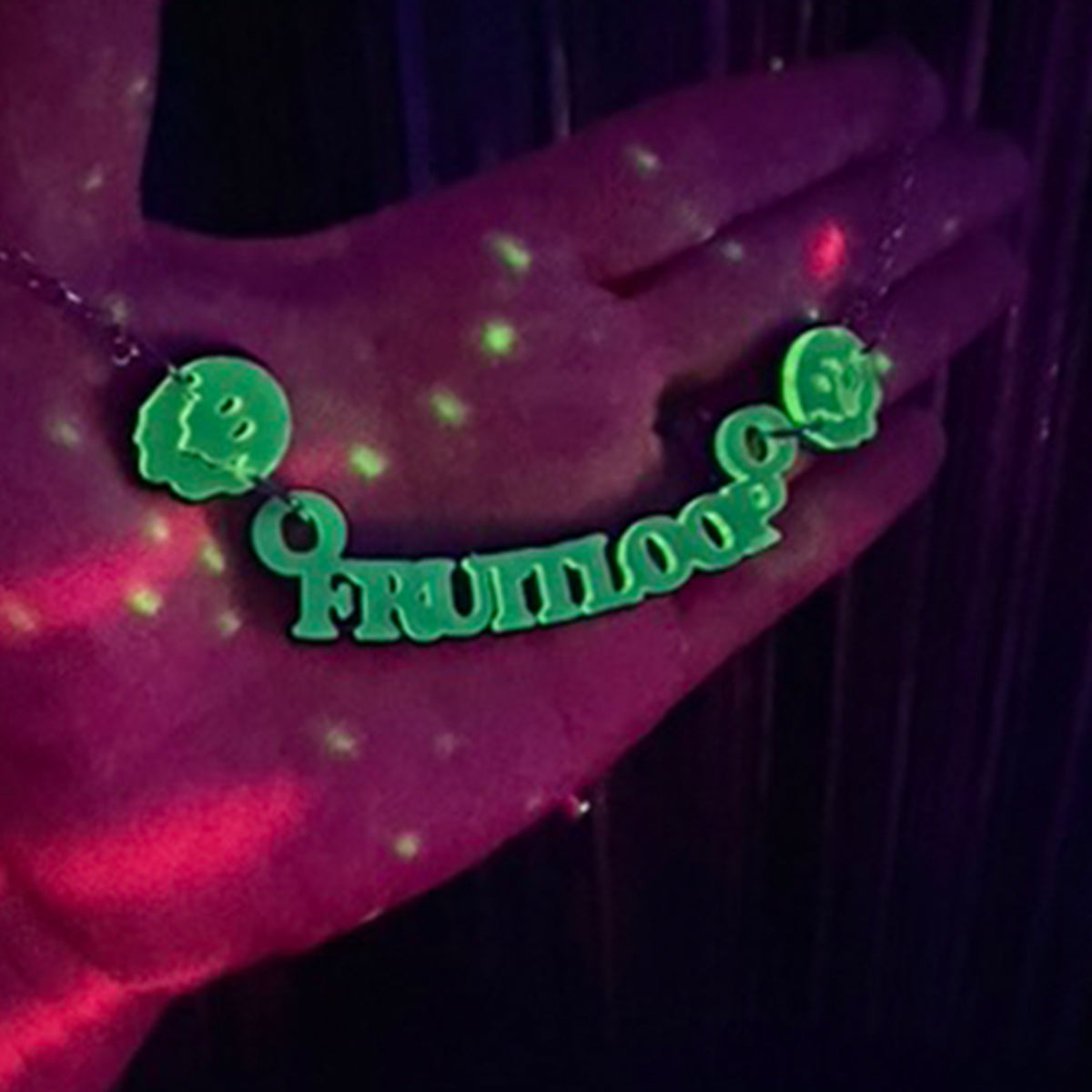 A blacklight-reactive necklace spelling 'fruitloop' with two green smiley faces, shown worn on a hand under UV lighting.