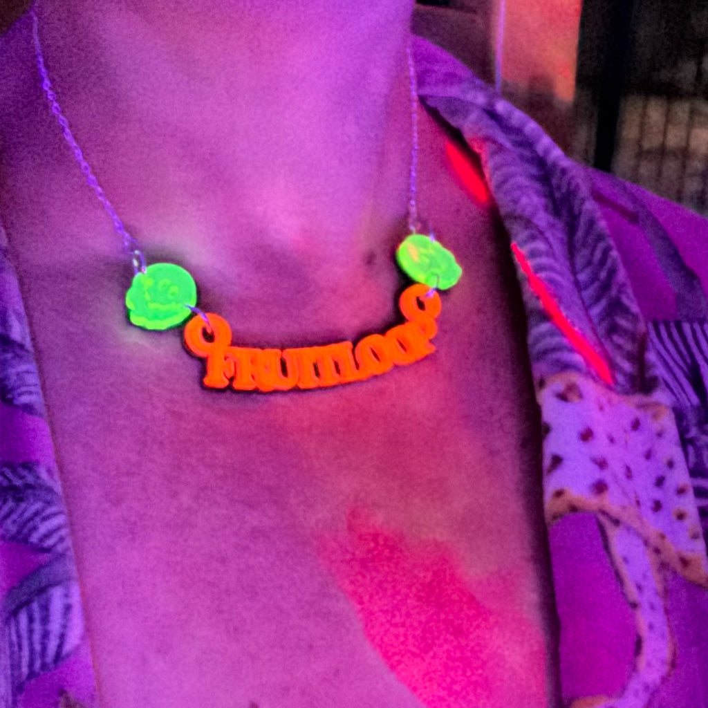 A close-up of a person wearing a necklace with a bright orange script word and two neon green smiley-face-like charms, shown under UV light with a purple cast.