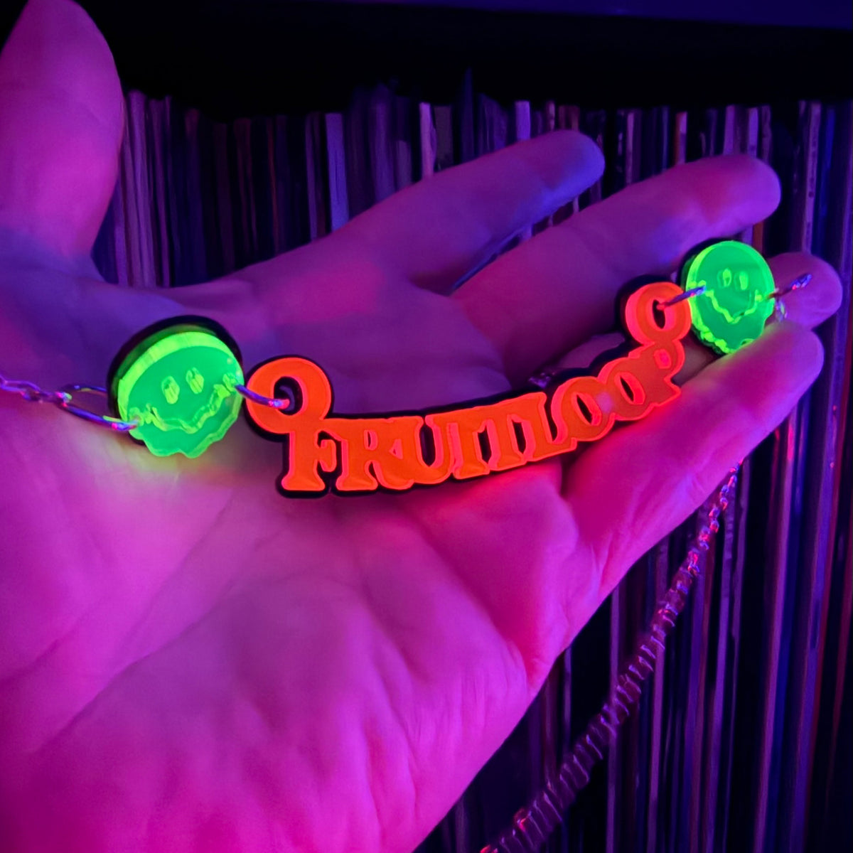 A neon-style necklace held in a hand under blacklight, featuring a red-orange script word 'FRUITLOOP' with two glowing green circular smiley-face charms on either side.