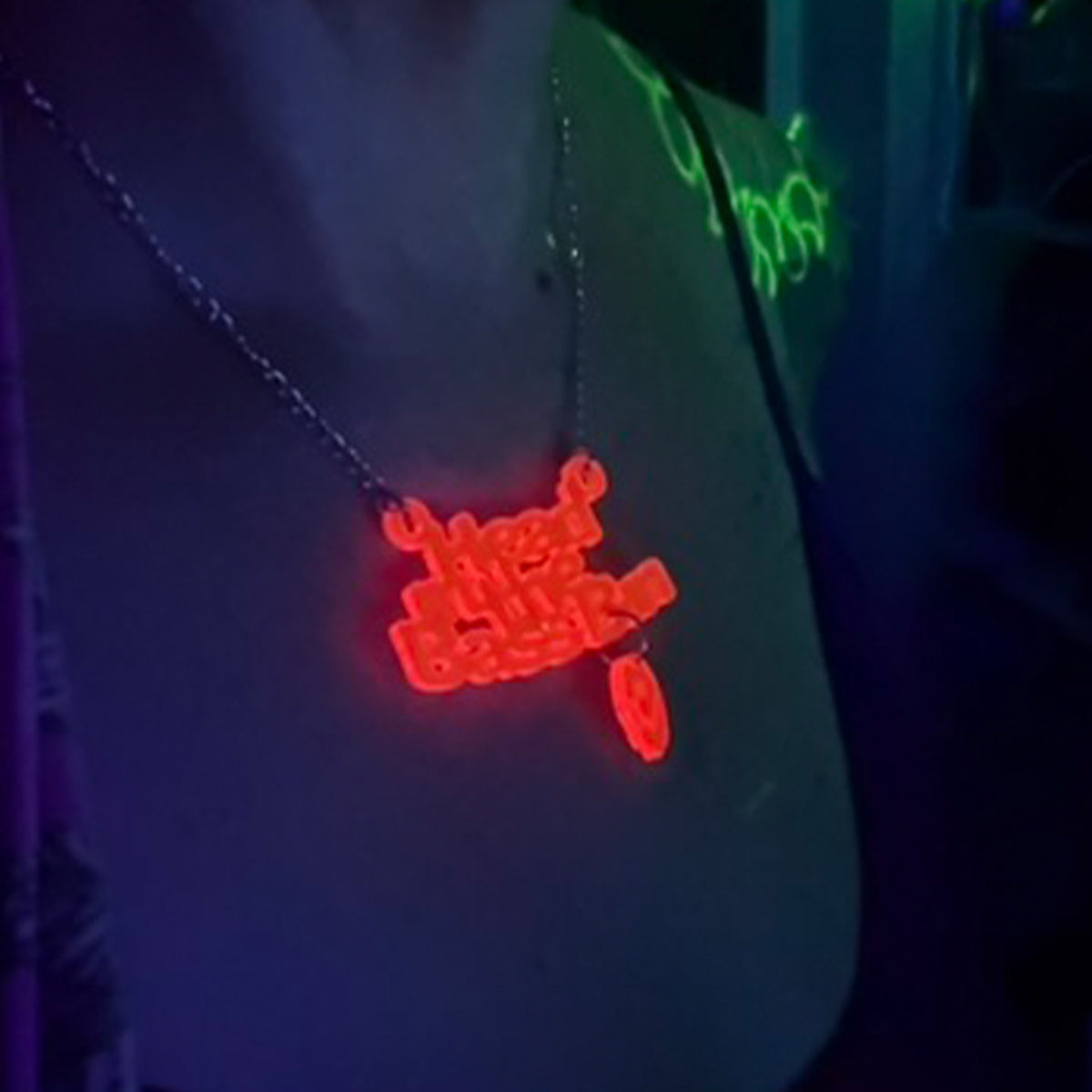 A glowing red neon-style pendant necklace on a chain, worn around the neck. The pendant appears to read 'head in the bass bin' in a blocky neon font, with a small matching red oval charm below it.