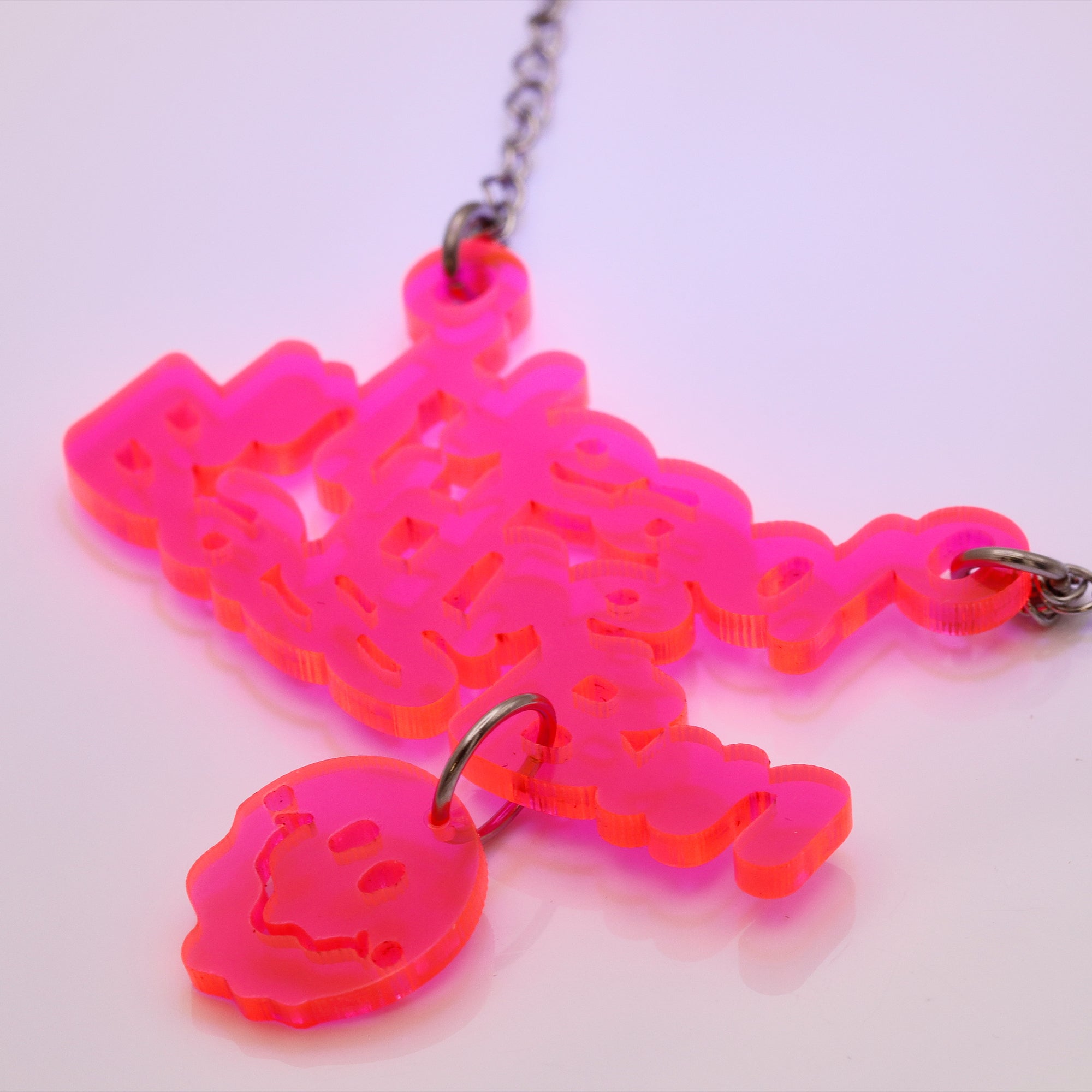 A neon pink acrylic pendant necklace spelling 'Head In The Bass Bin' with a matching small heart charm, shown on a silver chain against a light pink background.