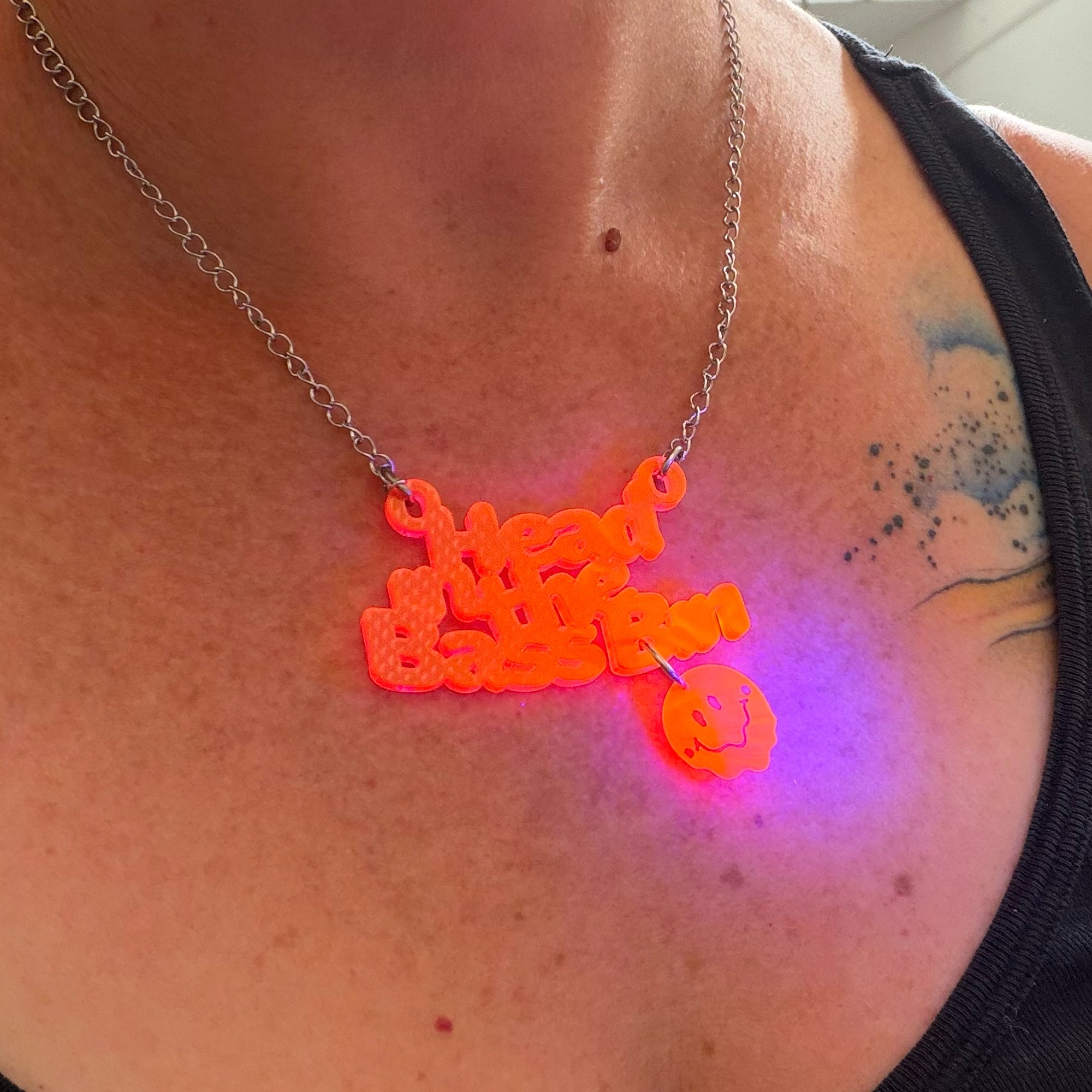 A close-up of a person's chest wearing a glowing orange 3D-printed necklace that reads 'Head in the bass bin' with a small acid skull charm, casting a purple light on the skin.