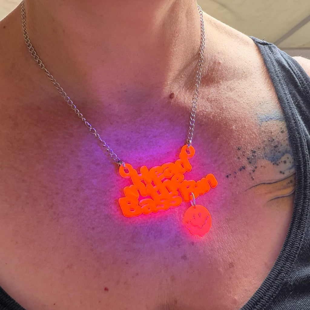 A close-up of a person's chest wearing a silver chain necklace with a glowing orange 'Head in the bass bin' pendant and a small matching glowing skull charm, under UV light that casts a purple glow on the skin.