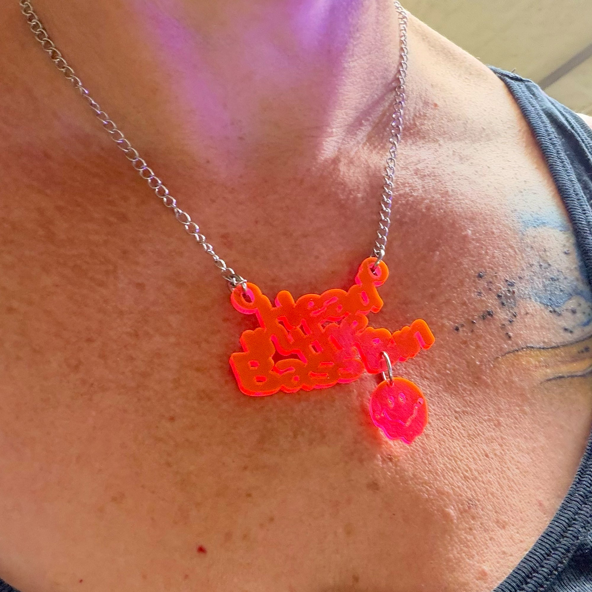A close-up of a person's neck and chest wearing a silver chain necklace with a bright neon orange-red 3D-printed pendant that reads 'head in the bass bin' and a small matching pink charm.