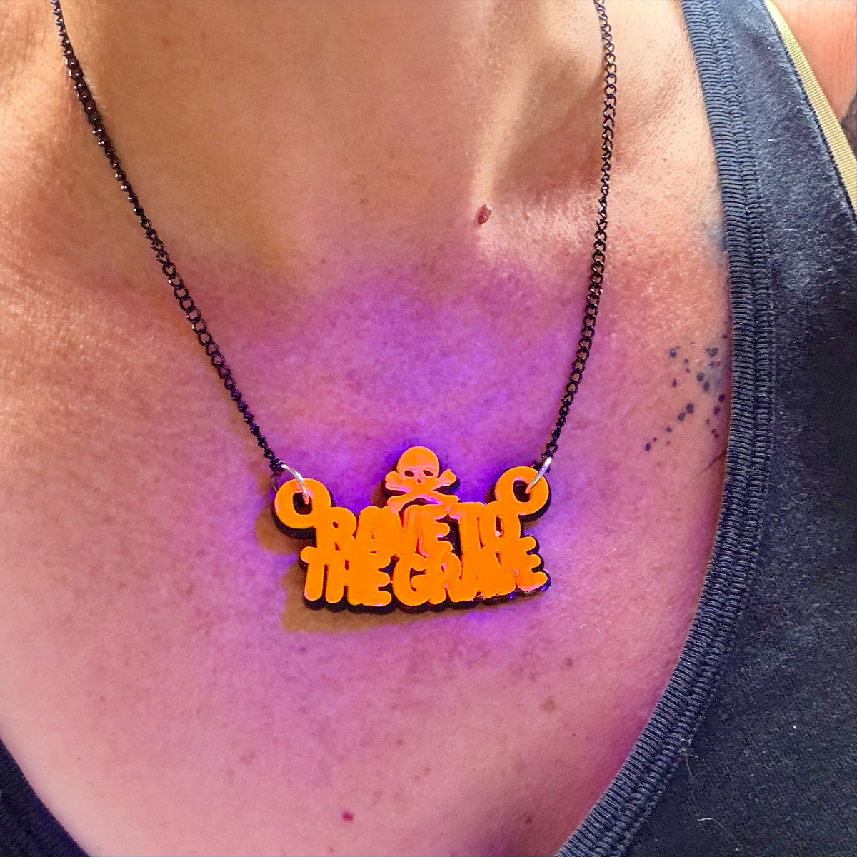 An orange acrylic pendant necklace with the word 'rave to the grave' and a small skull graphic, worn on a black chain over a person's chest under purple light.