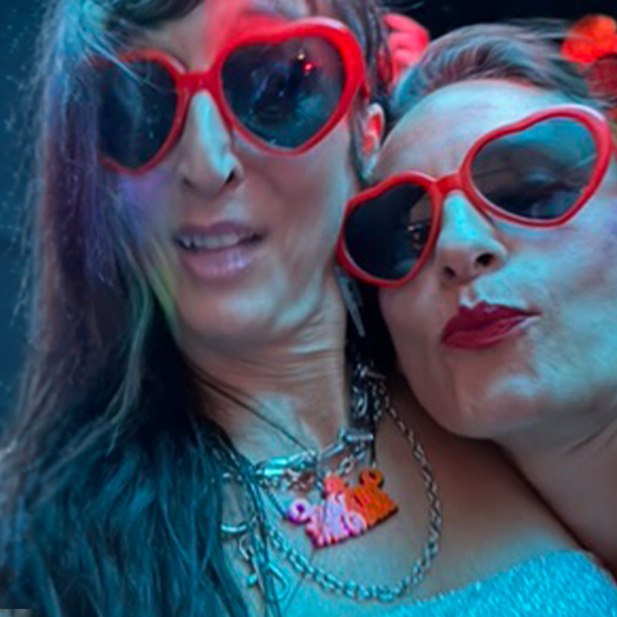 Two women wearing red heart-shaped sunglasses and layered silver necklaces, posing closely together under colorful red and blue lighting.