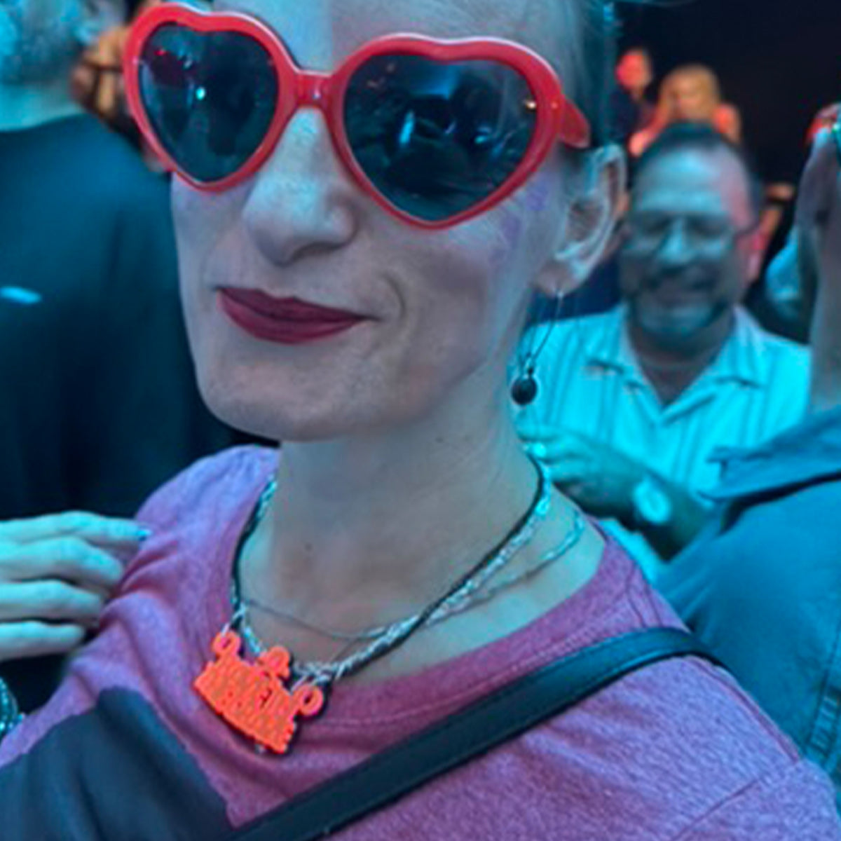 A person at a social event wearing bright red heart-shaped sunglasses and a pink top, with a colorful necklace in the foreground and blurred people in the background.
