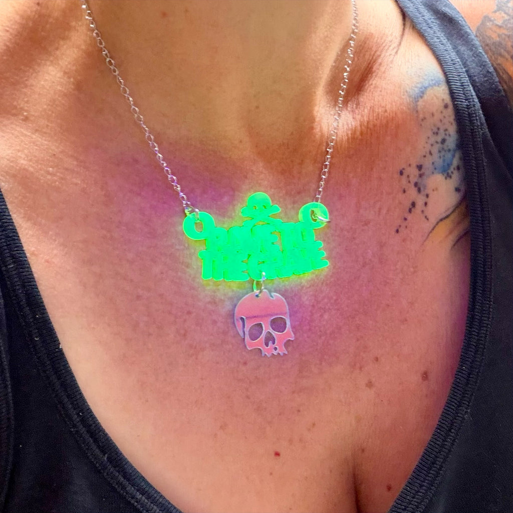 A close-up of a person's chest wearing a necklace that glows under UV light. The necklace features a silver-toned chain with a bright neon green nameplate spelling 'rave to the grave' and a dangling skull charm that shifts from pink to blue under light.