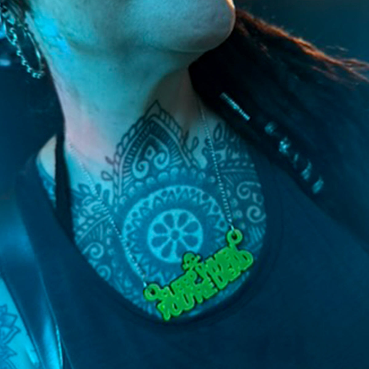 A close-up of a person's neck and chest wearing a silver chain necklace with a bright green script pendant spelling 'sleep when you're dead'. The scene is lit with a strong blue/cyan tint, highlighting the person's mandala tattoo on the neck.