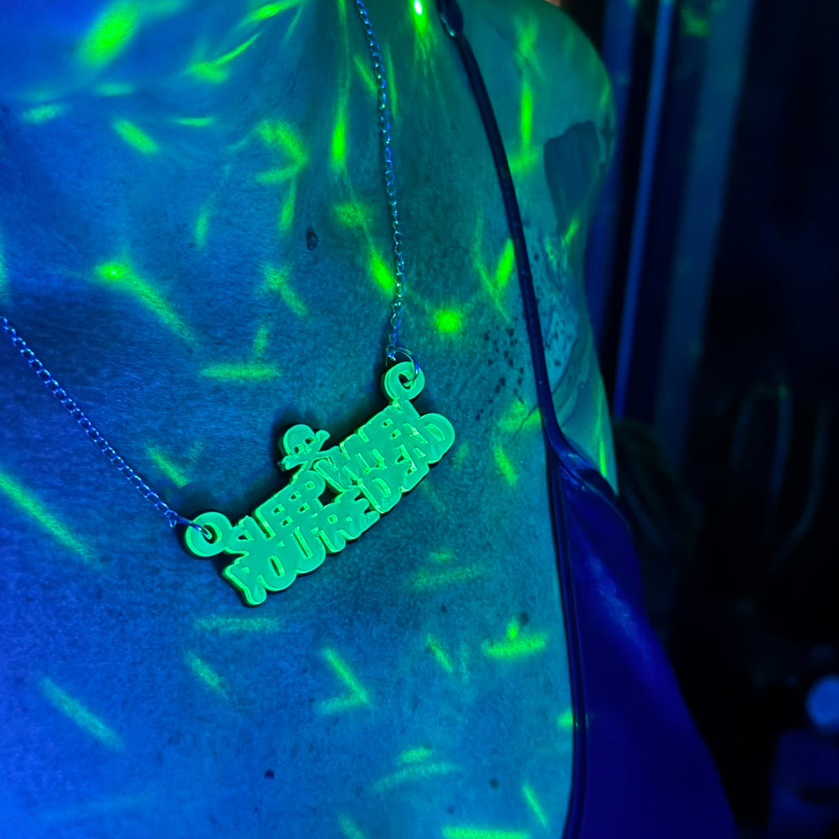 A close-up of a necklace with a stylized script pendant reading 'sleep when you're dead' on a chain, shown under UV light that casts a green glow on a blue surface.