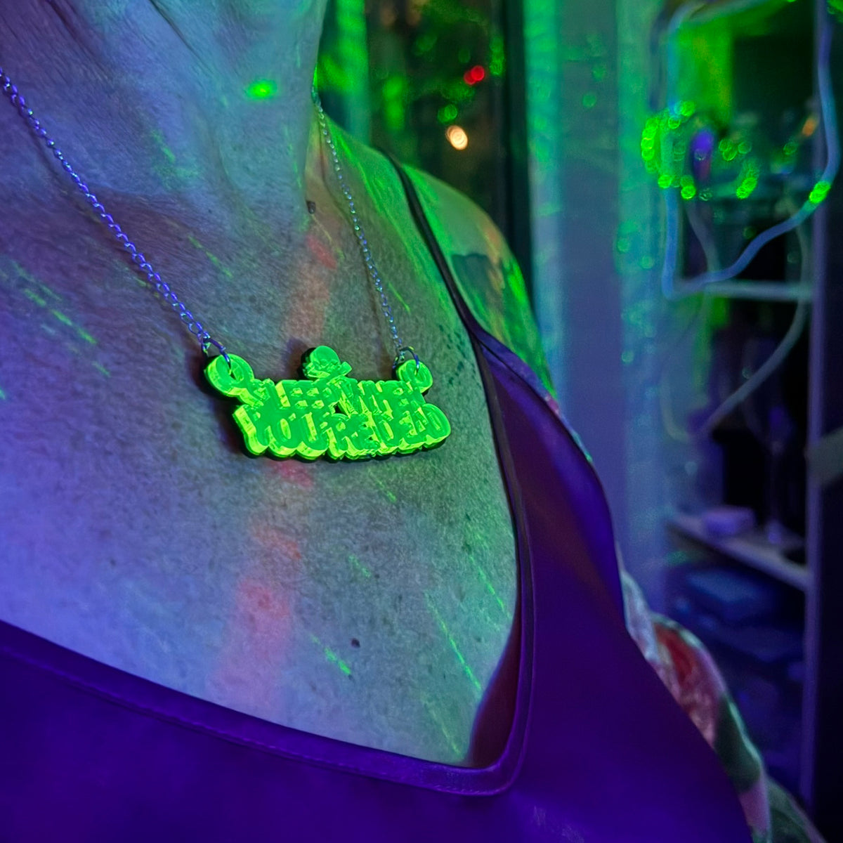 A person wearing a glowing neon green pendant necklace on a chain, photographed under blacklight in a dim room with green and purple ambient lighting.